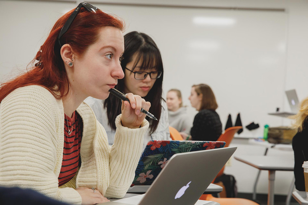 Two women in the foreground of a classroom. Both leaning over laptops and listening