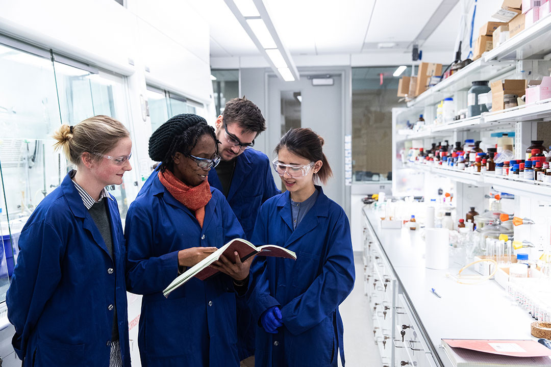 Four students in blue lab coats reviewing printed materials in a chemistry lab